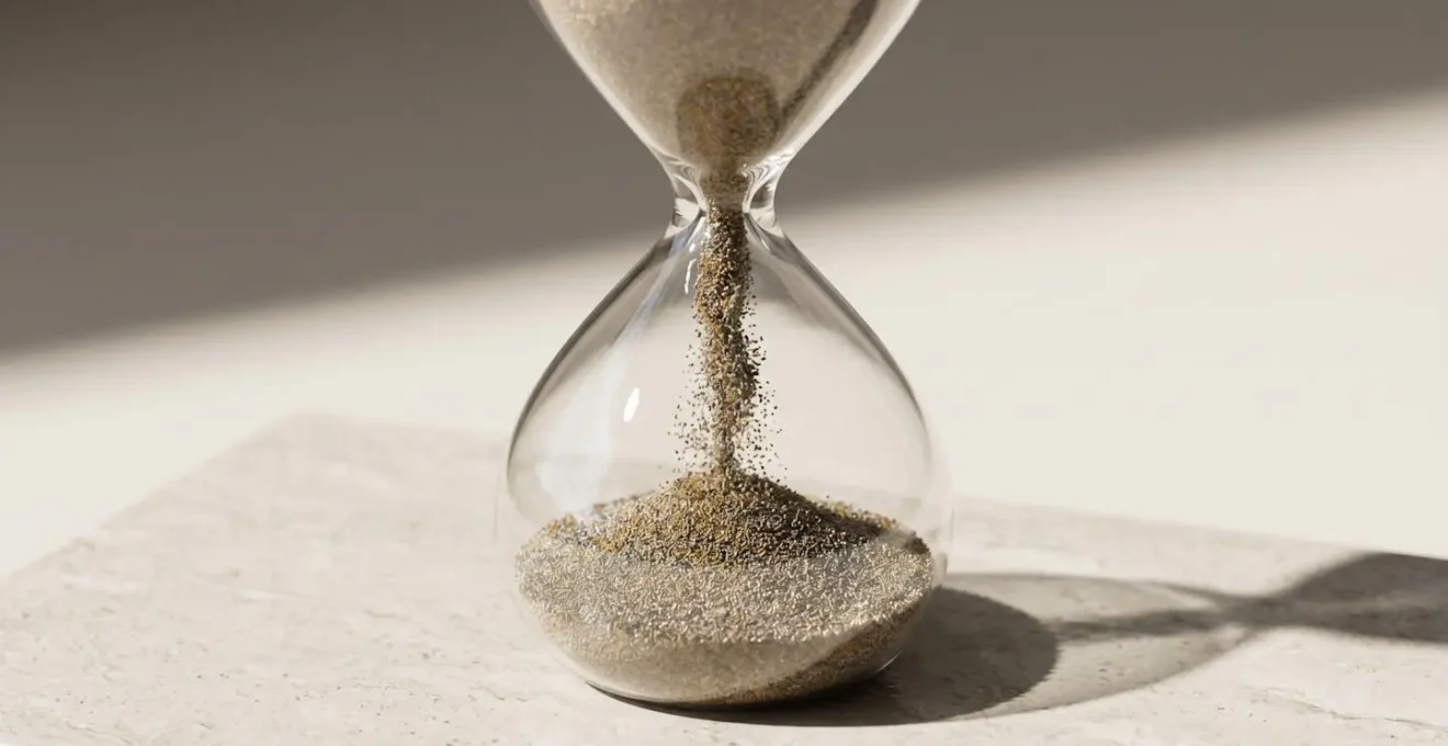 Sablier en verre dont l'écoulement du sable est ralenti par une accumulation de particules fines dans le passage central, photographié en lumière latérale sur fond neutre